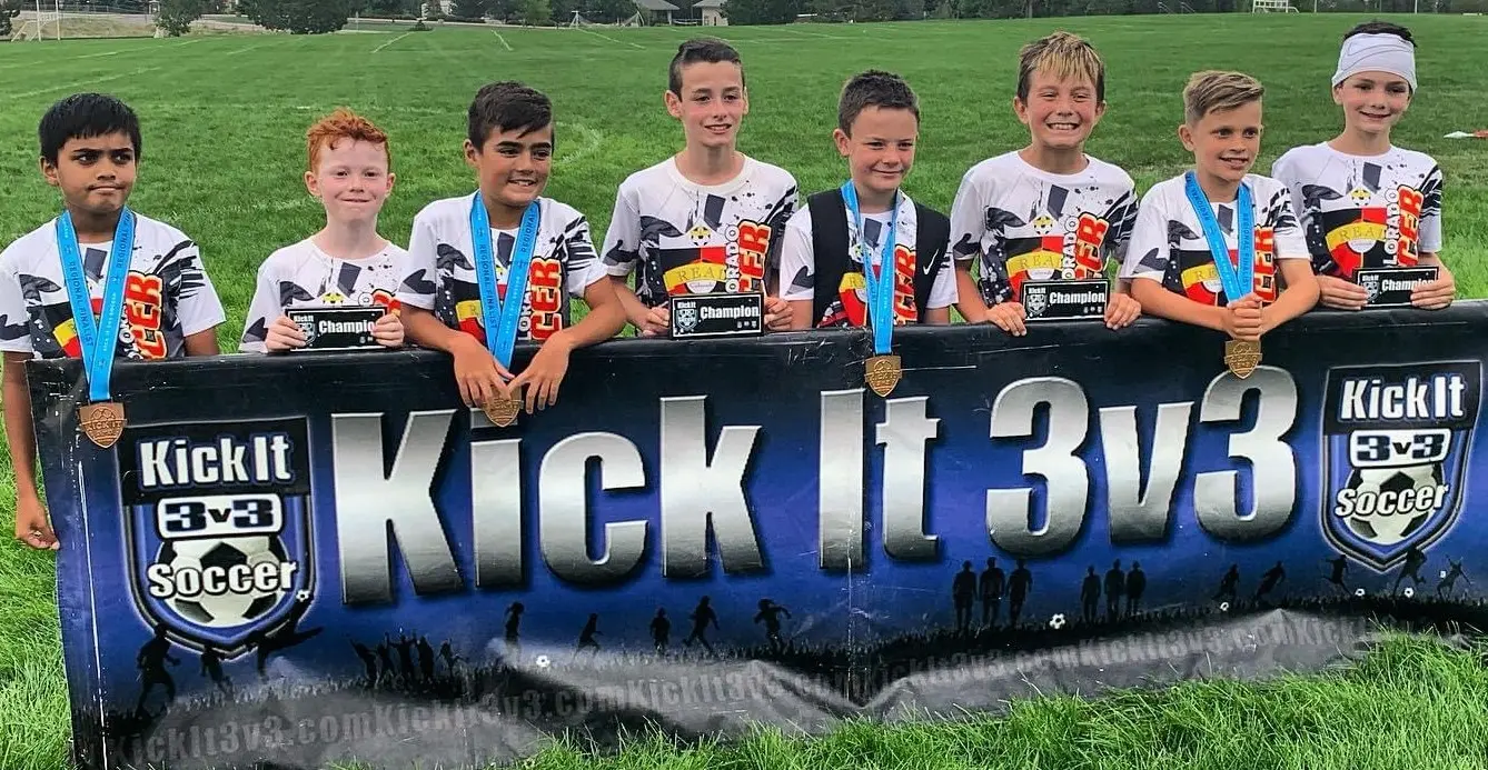 Kick It 3v3 Champions — 8 players with gold medals in front of Kick It 3v3 banner on grass field