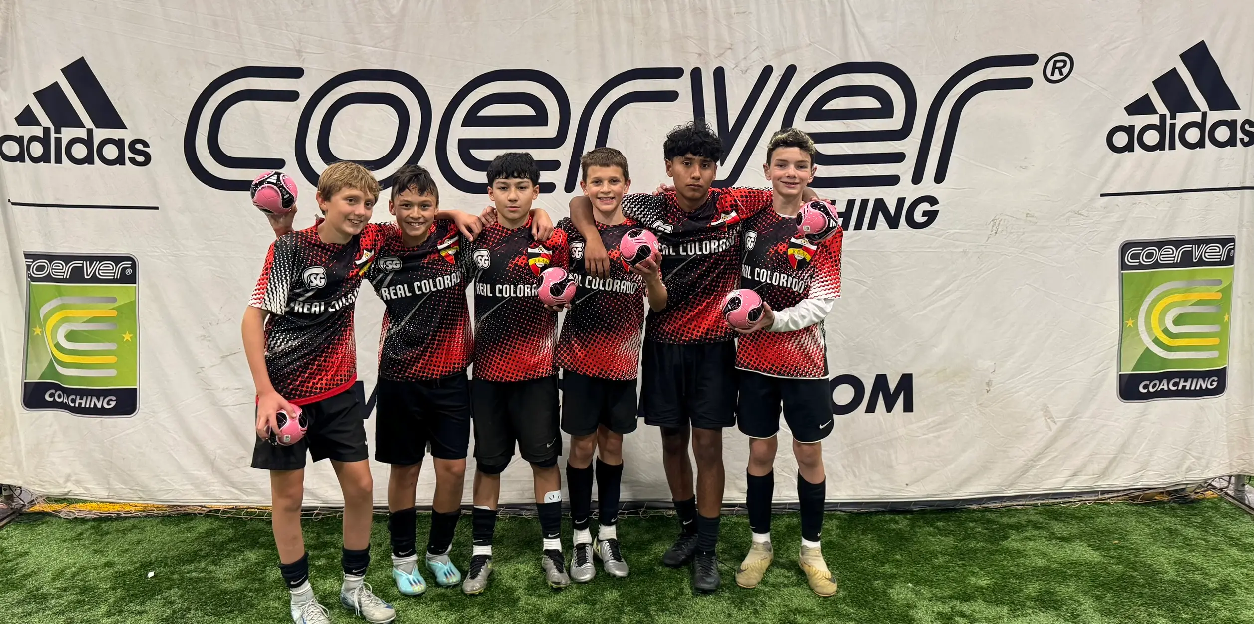 Real Colorado Academy team in black/red Coerver kits holding pink balls in front of Coerver Coaching adidas banner